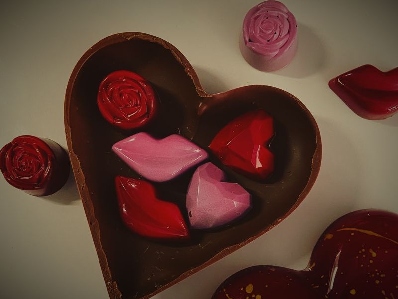 A chocolate heart box filled with lip, rose and heart chocolates, beside a red speckled chocolate heart